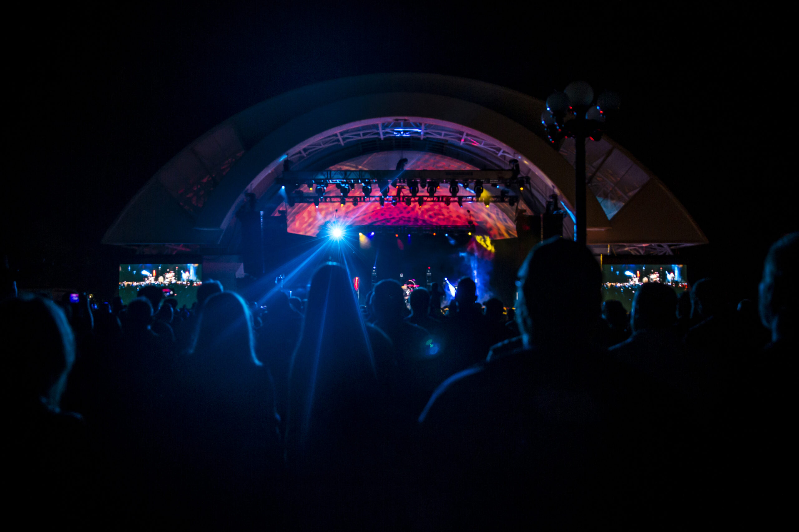 Image of the Bandshell at night. View of the crowd from behind as they enjoy a concert at night. The Bandshell stage is illuminated in blue and pink.