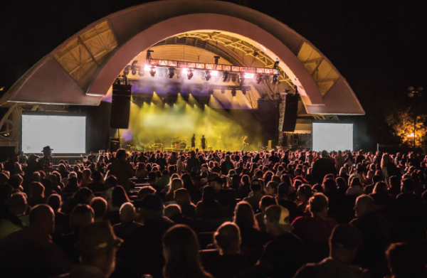 Image of the CNE Bandshell at night. The image includes a crowd of people listening to an artist perform on the stage.