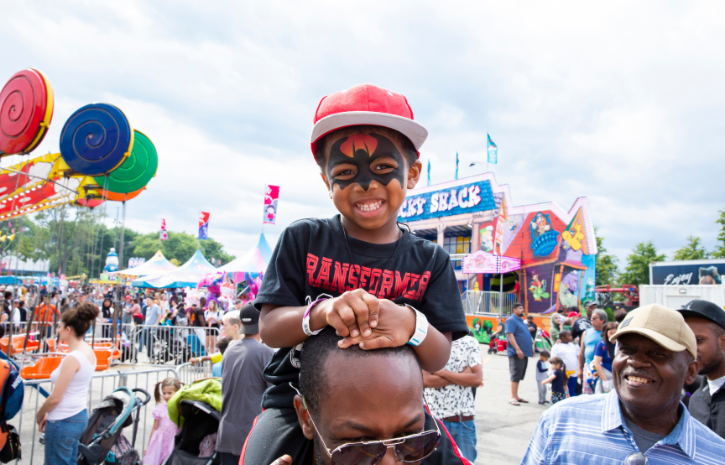 A photo of a boy wearing a baseball cap and face paint sitting on a mans shoulders